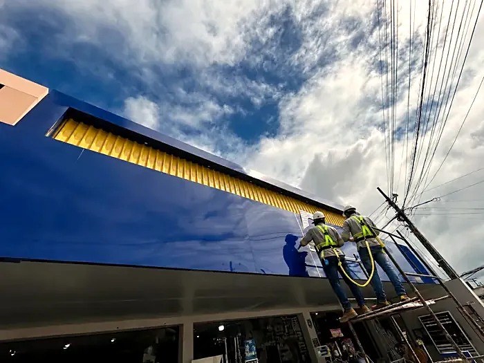 Dois instaladores da COMUNIKAR com capacete e cinto paraquedista aplicando o ACM azul na fachada