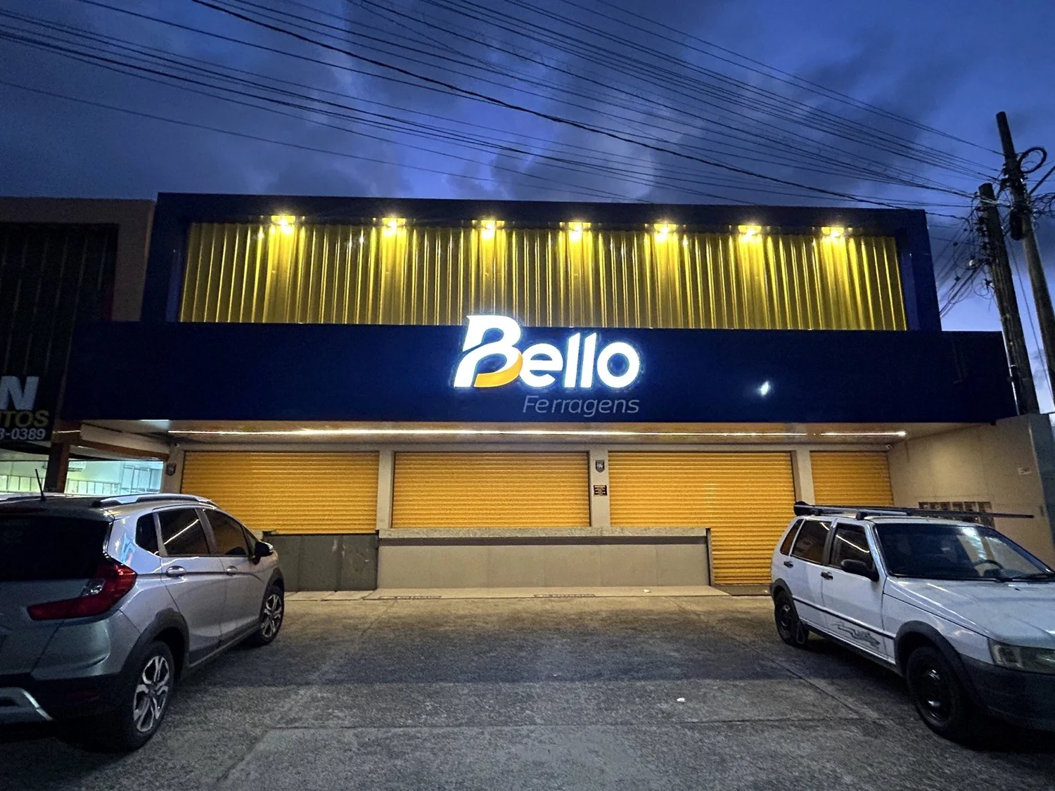 Fachada frontal completa acesa à noite, com letreiro branco sobre ACM azul e faixa dourada iluminada