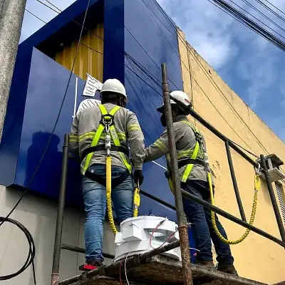 Dois instaladores da COMUNIKAR sobre andaime, de capacete e cinto paraquedista, montando o revestimento em ACM da fachada