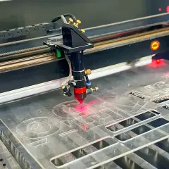 Cabeçote de corte a laser recortando letras em chapa de acrílico, com o feixe vermelho aceso sobre o material