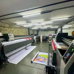 Galpão da fábrica com duas impressoras de grande formato e material impresso saindo para o chão